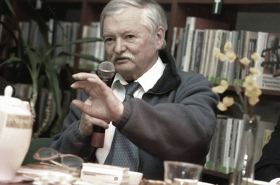 Poziome prostokątne kolorowe zdjęcie. Na nim siedząca przy stole postać mężczyzny (Z. Jerzyna ). W jednej ręce trzyma mikrofon, drugą ma wyciągniętą przed siebie.  W tle regał z książkami.Fot. Wanda Hansen