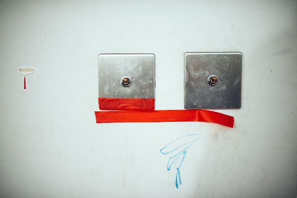 Pod włącznikami światła znajdują się dwie czerwone taśmy. Po lewej stronie widać naklejkę, a na niej narysowany pędzel malarski. Ściana na zdjęciu jest pomalowana kredkami. Fot. Zuzanna Jach
