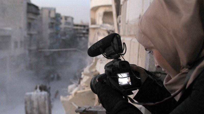 Obraz przedstawia kadr z filmu Dla Samy, na ujęciu widać syryjską dziennikarkę, filmującą z ukrycia zgliszcze miasta, które są widoczne w drugim planie przedstawianej sceny. Zdjęcie promuje pokaz filmu Dla Samy w cyklu Kino Kobiet.