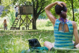 Na zdjęciu: kobieta siedząca tyłem do obiektywu podczas zajęć jogi w plenerze przy SDK; w tle drzewa i wieża obserwacyjna. Fot. Nel Lato