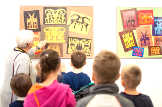 Zdjęcie przedstawia kadr z oprowadzania klasy szkolnej po Galerii Przy Kozach. Na zdjęciu znajduje się koordynatorka galerii wskazująca lewą ręką na dzieło bieżącej ekspozycji oraz grupa dzieci skierowana w stronę koordynatorki.
