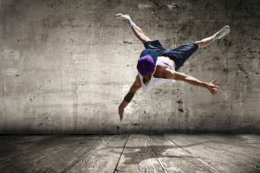 Zdjęcie przedstawia tancerza hip-hop wykonującego figurę taneczną. W tle betonowa przestrzeń.