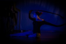 Na zdjęciu widoczna jest tancerka siedząca na scenie z rękami podniesionymi do góry. Na scenie przeważa niebieskie, ciemne światło. 