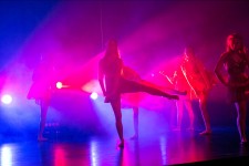 Zdjęcie przedstawia grupę tancerek w trakcie ruchu na scenie. Na scenie przeważa światło niebieskie oraz czerwone. 