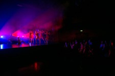 Na zdjęciu widoczna jest scena z tancerkami oraz fragment publiczności. Wszystko w ciemnych kolorach czerwieni i niebieskiego. 
