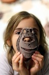 Na zdjęciu widzimy dziewczynkę, która przykłada do twarzy maskę wykonaną z gliny.