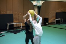 Na zdjęciu widoczne są dwie dziewczynki na macie do ćwiczeń, które podają sobie piłkę nad głowami stojąc do siebie tyłem. 