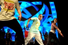 Zdjęcie przedstawia grupę taneczną w trakcie pokazu na scenie. Na pierwszym  planie widać trzy postacie w tanecznej pozie. 
