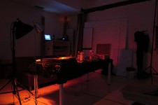 Na zdjęciu studio fotograficzne z przyciemnionym, czerwonym światłem. Na środku studia znajduje się stół oraz kilka lamp studyjnych. 