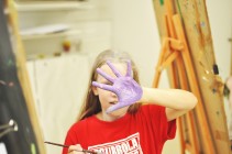 Na zdjęciu: dziewczynka przy sztalugach zakrywająca twarz pomalowaną dłonią. Fot. SDK
