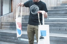 Na zdjęciu mężczyzna trzymający w rękach materiałowe torby ozdobione na warsztatach sitodruku. Mężczyzna stoi na drewnianym amfiteatrze SDK.