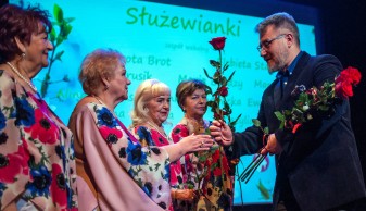 Na zdjęciu 4 członkinie zepołu wokalnego otrzymują róże od Burmistrza Rafała Miastowskiego. Burmisrz jest ubrany na ciemnon, wokalistki są ubrane w kwieciste bluzki. W tle grafika z wymienionymi nazwiskami wokalistek, wymienione w artykule.