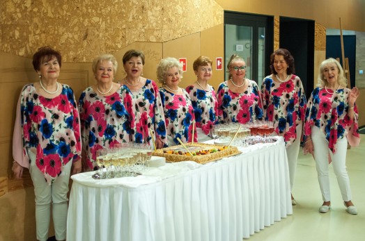 Na zdjęciu osiem pań - wokalistek przed urodzinowym tortem ustawionym na białym podłużnym stole. Paniew ubrane są w białe spodnie i kwieciste bluzki. W tle korkowa ściana.