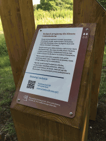 Zdjęcie prezentuje tabliczkę informacyjną ścieżki. W tle widoczny Park Służewskiego Domu Kultury.