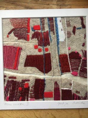 Zdjęcie pzredstawia pracę - mieszaninę technik z zastosowaniem tkaninowych aplikacji i haftu w wielu odcienach czerwieni. Obraz przedstawia plan miasta na lnianym tle. Obramowanie prac białe.