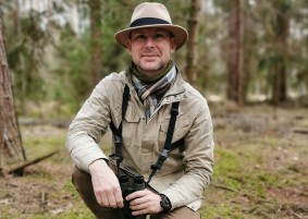 Na zdjęciu: Adam Zbyrt w lesie, ubrany w kapelusz, w dłoniach trzyma lornetkę. Przyklęka na jedno kolano. Fot. zasoby Adama Zbyrta