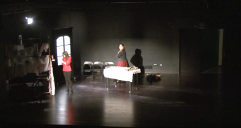 Kadr z filmu. Na scenie teatralnej stoją dwie kobiety. Scenografia przypomina pokój, z lewej strony znajdują się drzwi, a na środku sceny stoi stół przykryty obrusem. 