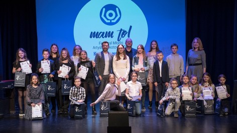 Na zdjęciu: grupa osób na scenie na tle ekranu z logo Konkursu Wokalnego MAM to! 2022; z przodu kuzające dzieci z dyplomami laureatów, za nimi stojący jurorzy i inne dzieci z dyplomami.