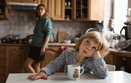 Na zdjęciu, na pierwszym planie widać 5 letnią dziewczynkę przy stole, opierającą głowę rączką i pijącą kakao przez słomkę z kubka. W tle w kuchni widnieje postać kobiety. fot. Zuzanna Jach