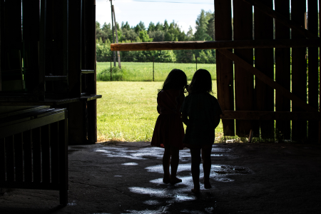 Na zdjęciu widać zarys dwóch kilkuletnich dziewczynek stojących tyłem do aparatu w stodole i patrzących w stronę łąki. Fot. Zuzanna Jach