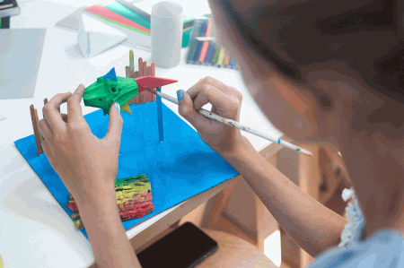 Na zdjęciu widoczne ręce młodej uczestniczki warsztatów plastycznych oraz wykonywana przez nią praca - kolorowa ryba pomalowana farbami. Praca trzymana jest nad kartką papieru pomalowaną na niebiesko jako morze. fot. Dominika Dzieniszewska 