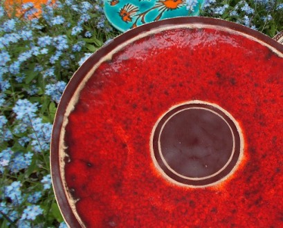 Na zdjęciu praca ceramiczna Moniki Marks WItkowkiej sfotografowana od góry. Patera jest krwistoczerwowa z obramowaniem w odcieniach brązu. Praca ceramiczna jest uożona na kwiecistej łące.