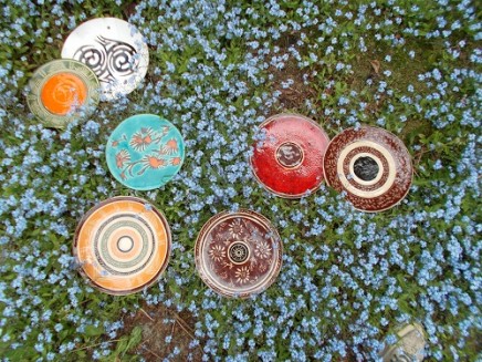 Na zdjęciu prace ceramiczne Moniki Marks WItkowkiej sfotografowane od góry. Patery są wielobrawne, domunuje w nich brąz, czerwień, żółć z kołowymi obramowaniami i elementami ludowymi. Prace ceramiczna są ułożone na kwiecistej łące.