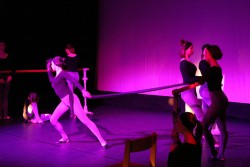  Mocne, czerwone światło. Grupa tancerek przy drążkach baletowych ustawionych po obu stronach sceny. W centralnym punkcie widać dynamicznie poruszającą się tancerkę z długimi, elastycznymi rękawami, rozciągniętymi do obu drążków.