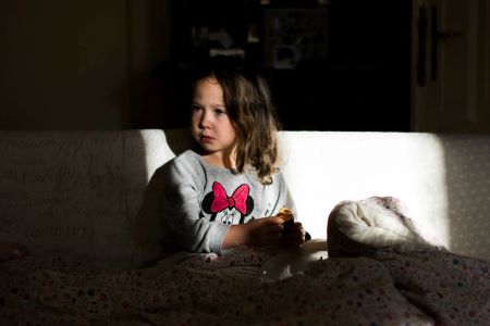 Na kanapie siedzi dziewczynka trzymająca w rękach racucha. Patrzy się w prawą stronę ze zdumioną miną. Jest przykryta kołdrą, a na jej twarz pada światło z okna. Fot. Zuzanna Jach