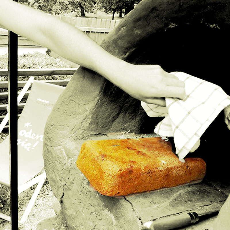 Zdjęcie: Fragment glinianego pieca chlebowego w ogrodzie SDK. Widoczne kobiece ramię, ze ściereczką w dłoni wyciągające z niego upieczony prostokątny chleb. Fot. Dominika dzieniszewska