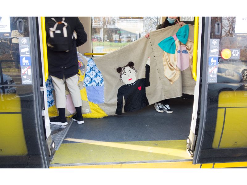 Zdjęcie przedstawia fragment instalacji w trakcie przejazdu tramwajem - autoportret wolontariuszki, która macha do obiektywu aparatu.