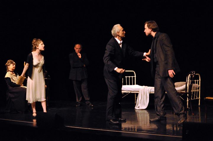 Zdjęcie wykonane w trakcie przedstawienia. Na zdjęciu aktorzy na scenie teatralnej. Mężczyzna na pierwszym rozmawia z innym mężczyzną. Obok stoi kobieta. W tle pozostali aktorzy. 
