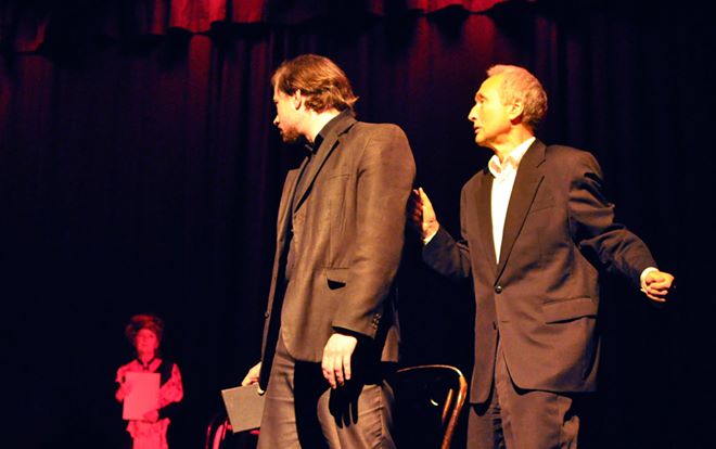 Zdjęcie wykonane w trakcie przedstawienia. Na zdjęciu aktorzy na scenie teatralnej. Dwóch mężczyzn na pierwszym planie rozmawia ze sobą. 