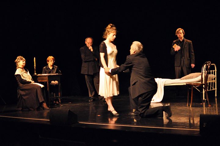 Zdjęcie wykonane w trakcie przedstawienia. Na zdjęciu aktorzy na scenie teatralnej. Mężczyzna na pierwszym planie klęczy przed kobietą. W tle pozostali aktorzy. 